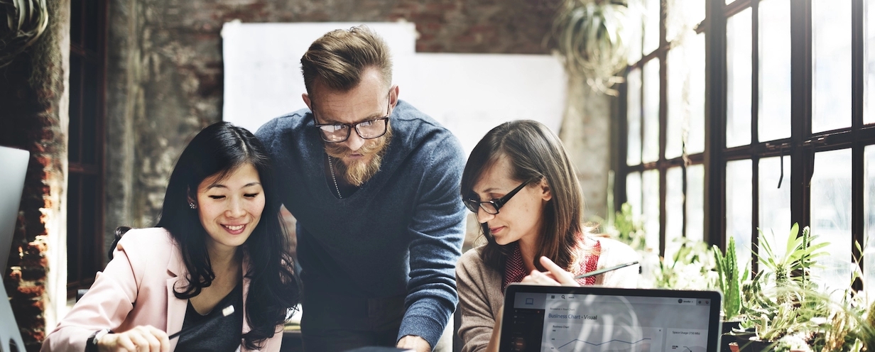 Photo of 3 people huddled around a desk looking at a notebook with open laptop in foreground