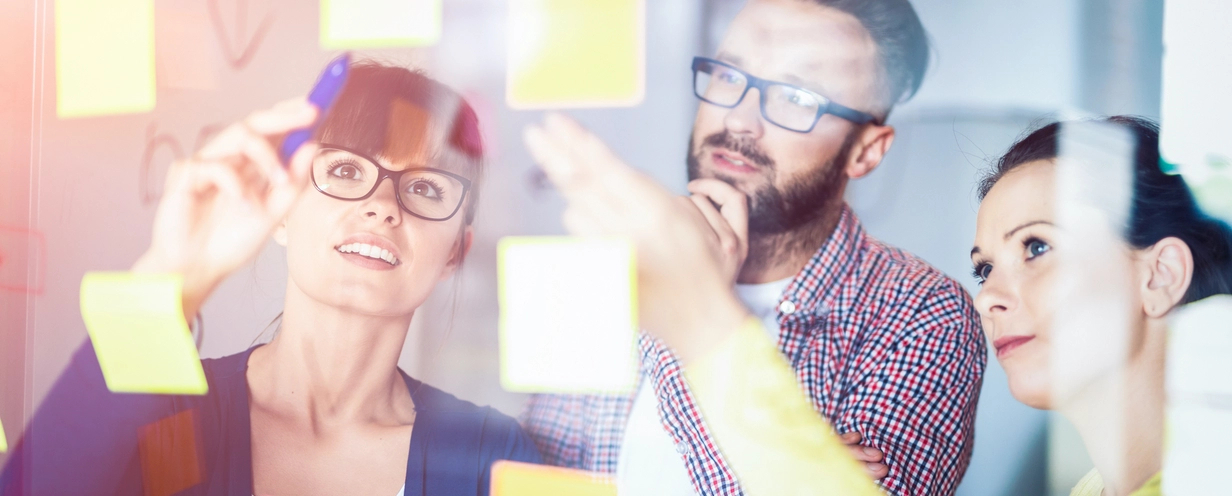 Photo of 3 people adding sticky notes to a clear board, shown from the other side of the board