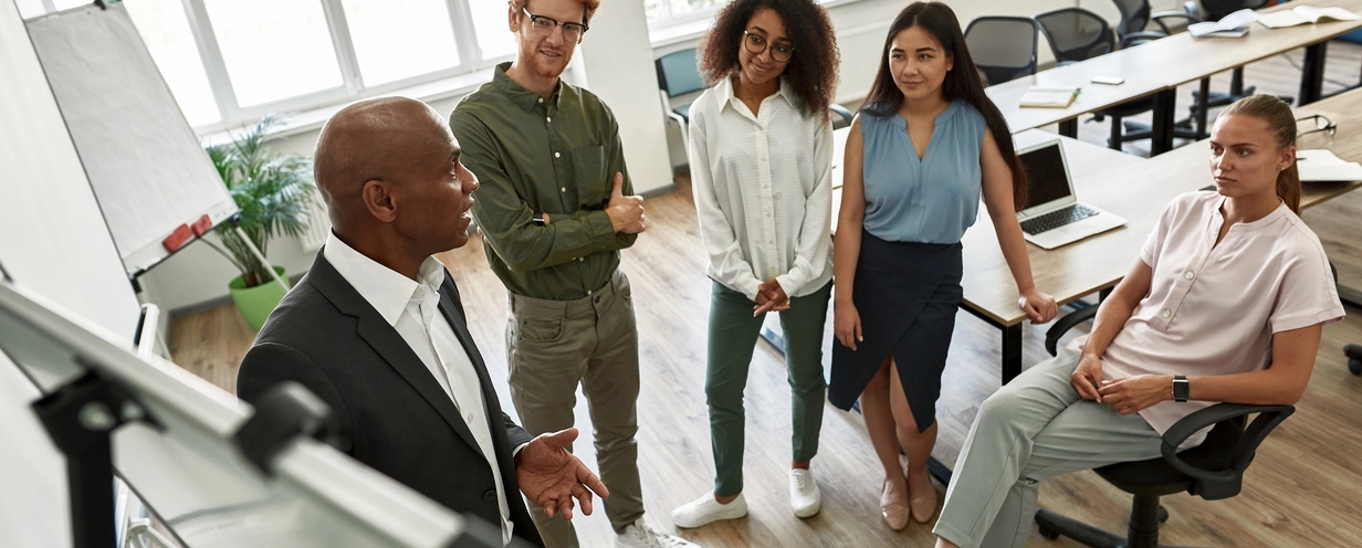 A diverse group of colleagues standing in a modern office, attentively listening to a presentation given by a man in a suit at a whiteboard