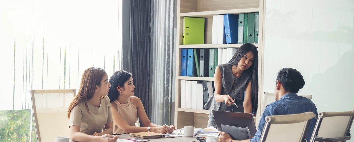 Group of colleagues in a meeting room, with one woman standing and pointing at a laptop screen while the others listen attentively