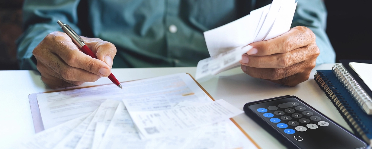 Close-up of a person’s hands holding receipts and writing on a document with a pen, while a smartphone with a calculator app open