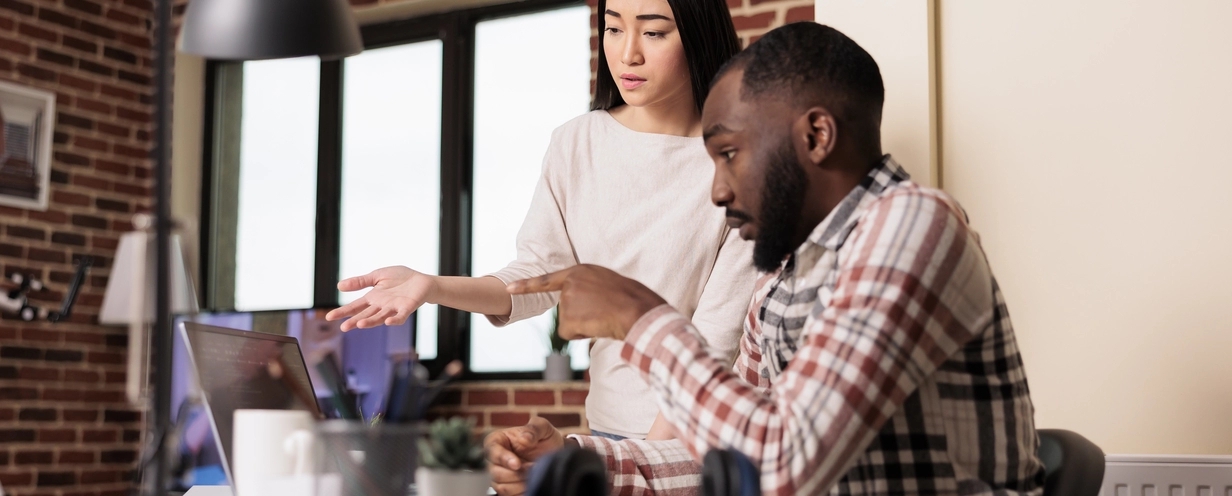 Photo of a man and a woman at a desk pointing at a computer screen having a discussion