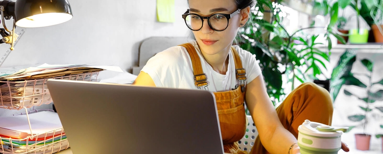 Photo of a woman wearing glasses drinking coffee sitting at desk with laptop