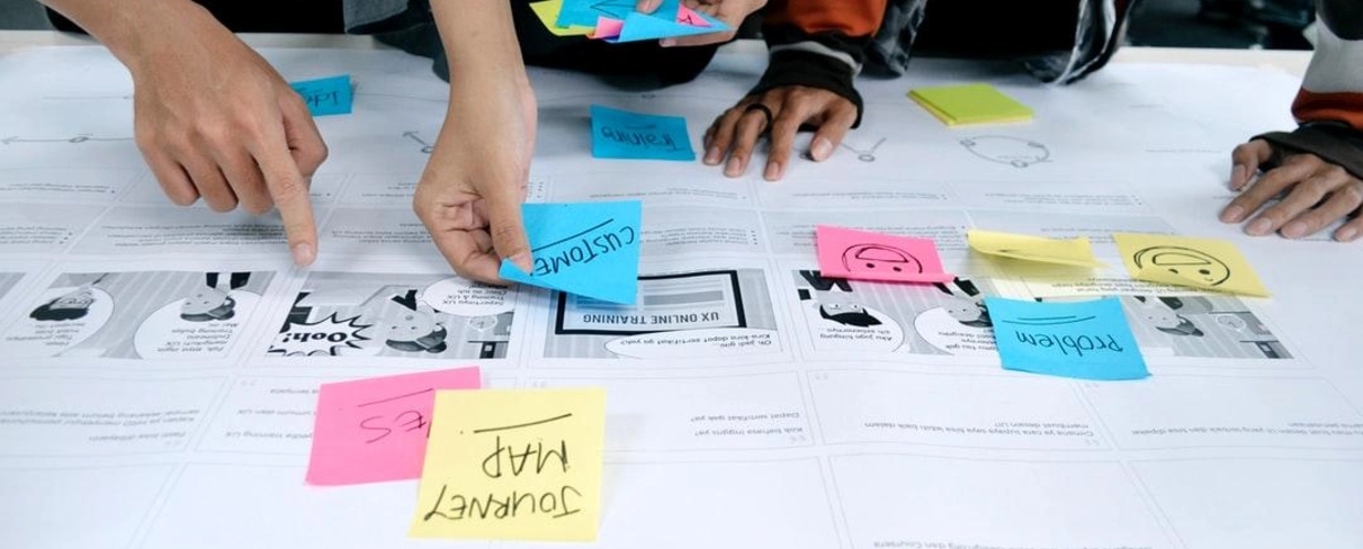 photo of the hands of multiple people adding post-it notes to a large sheet of paper on a conference table