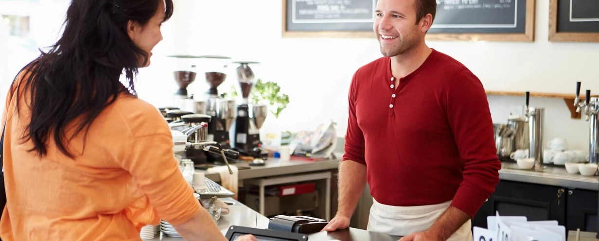 Photo of someone checking out at a coffeeshop using a laptop as a barista smiles