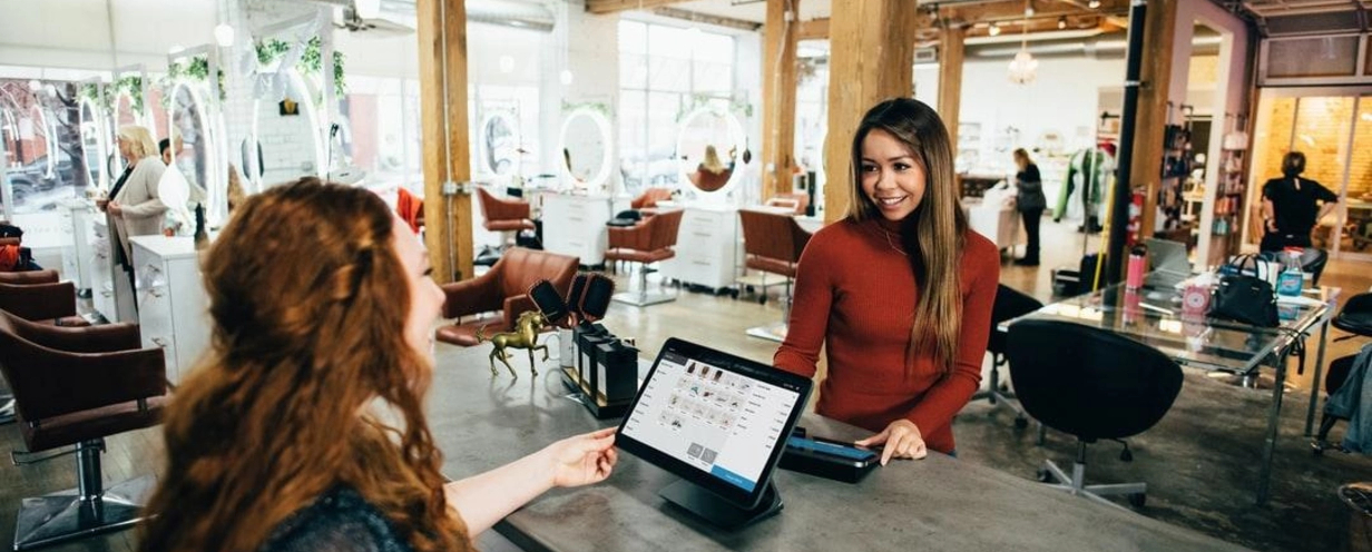 A woman at a salon checkout counter smiling as she interacts with a client, while using a touchscreen point-of-sale system