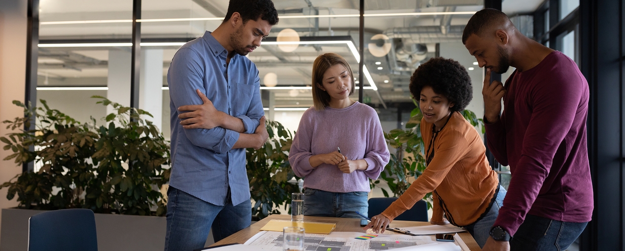 4 individuals in an office surrounding a table and looking at work documents.