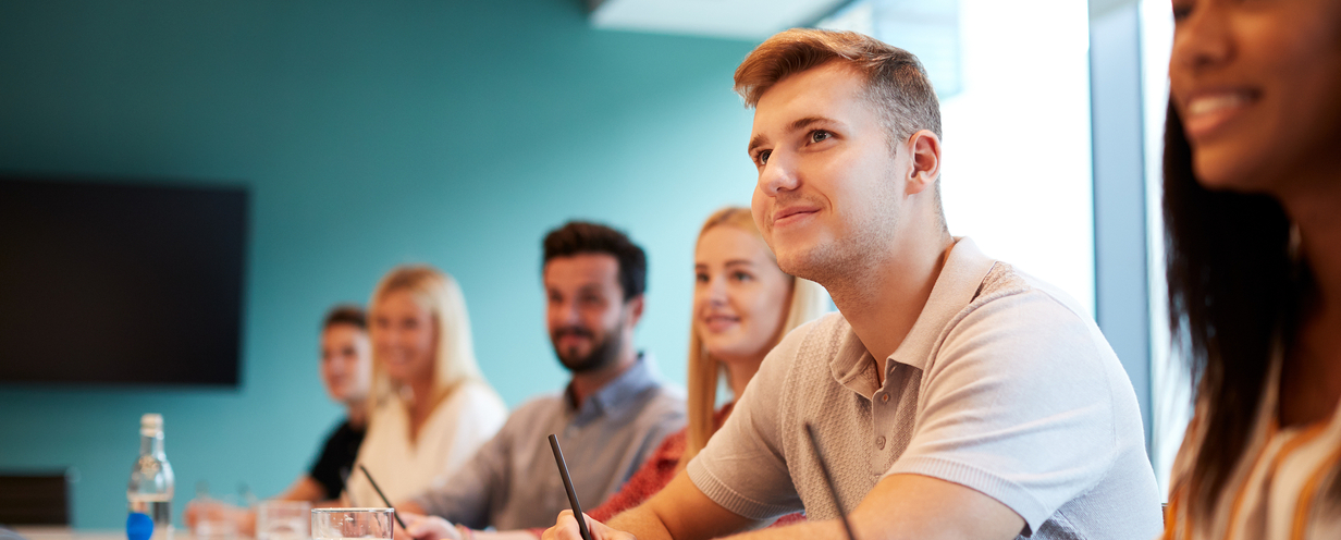 Group of young individuals sitting at a boardroom table watching a speaker present