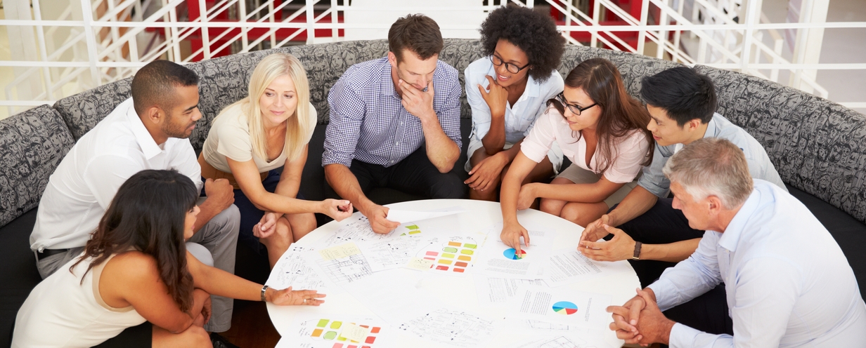 Photo of 7 professional colleagues seated around a circular coffee table looking at marketing campaign