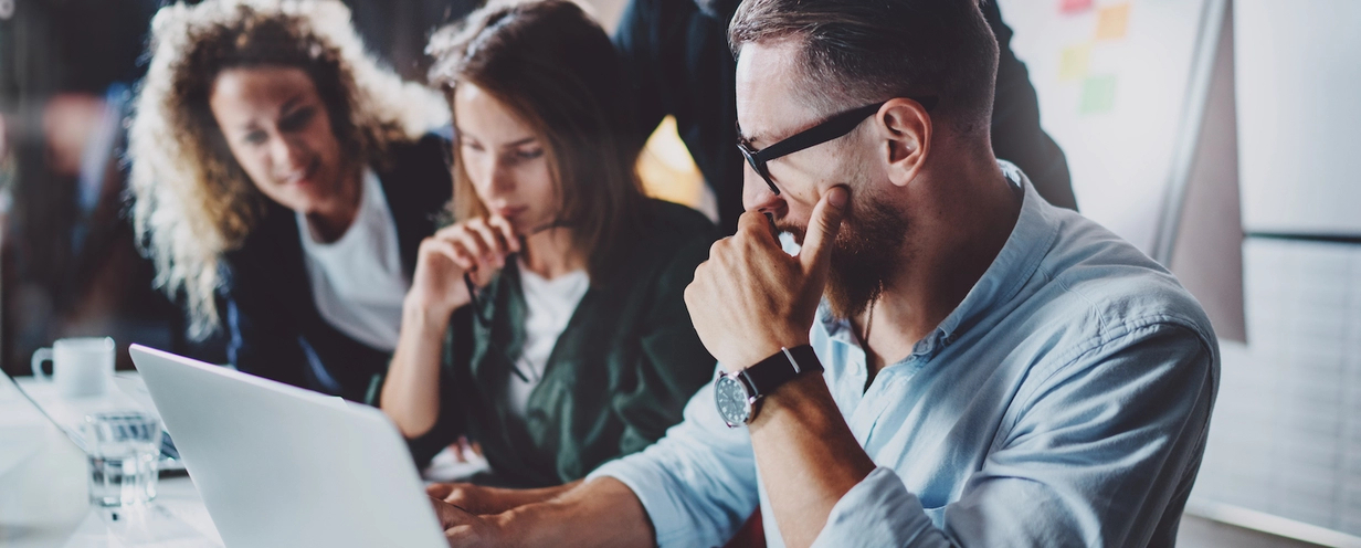 Focused group of colleagues collaborating at a desk, with a man in glasses intently working on a laptop while two women observe and discuss