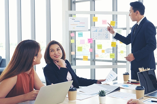 Three people in a meeting room, with one person presenting a board covered in colorful sticky notes, illustrating a project plan aligned with SAFe Lean Agile principles.