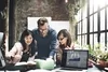 Photo of 3 people huddled around a desk looking at a notebook with open laptop in foreground