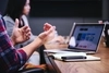 Photo of two people sitting near a laptop. One person is gesturing with their hands. 