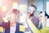 Photo of 3 people adding sticky notes to a clear board, shown from the other side of the board