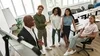 A diverse group of colleagues standing in a modern office, attentively listening to a presentation given by a man in a suit at a whiteboard