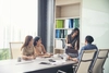 Group of colleagues in a meeting room, with one woman standing and pointing at a laptop screen while the others listen attentively