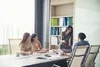 Group of colleagues in a meeting room, with one woman standing and pointing at a laptop screen while the others listen attentively