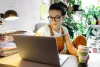 Photo of a woman wearing glasses drinking coffee sitting at desk with laptop