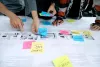 photo of the hands of multiple people adding post-it notes to a large sheet of paper on a conference table