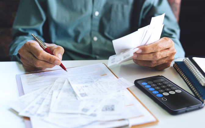 Close-up of a person’s hands holding receipts and writing on a document with a pen, while a smartphone with a calculator app open