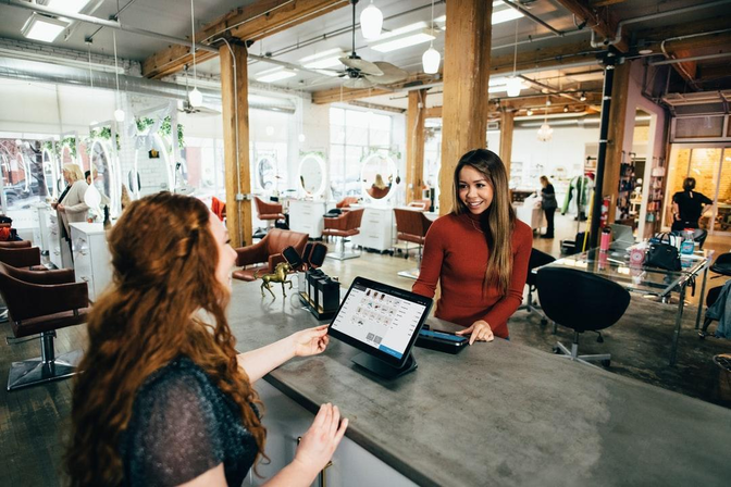 A woman at a salon checkout counter smiling as she interacts with a client, while using a touchscreen point-of-sale system