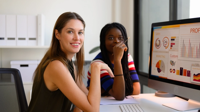 Photo of two women sitting at a desk looking at monitor showing a dashboard of ecommerce metrics