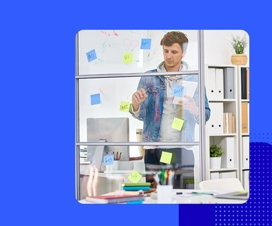 Man standing at desk writing on a clear board, viewed from the opposite side